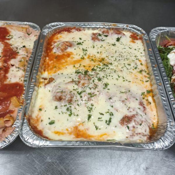 Three foil trays of Italian food, including baked ziti and lasagna with melted cheese and parsley.