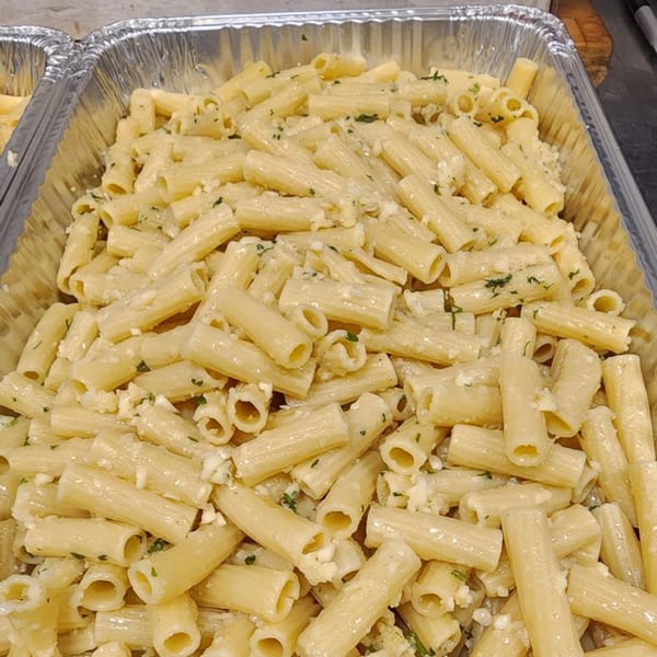 A large aluminum tray filled with rigatoni pasta in a creamy white sauce, garnished with herbs.