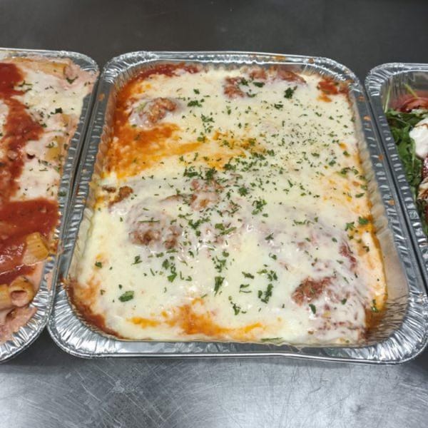 Three aluminum trays of Italian food, including baked ziti, lasagna, and a salad.