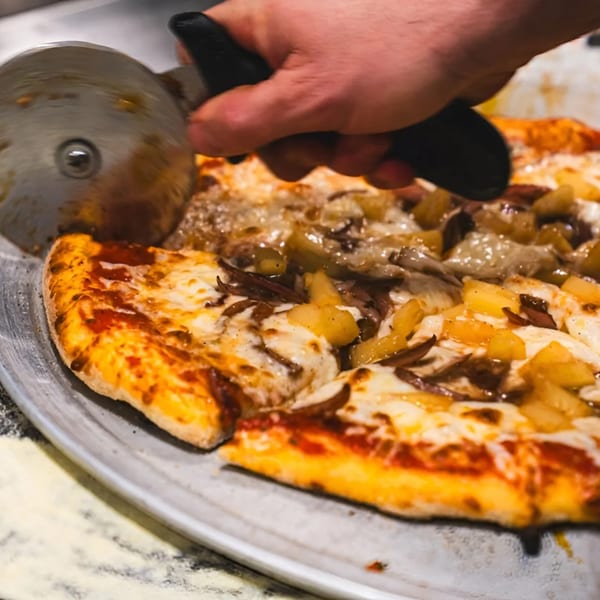 A hand uses a pizza cutter to slice a Stallone's Italian Eatery pizza topped with pineapple and onions.